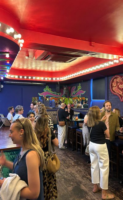 A bustling, brightly decorated bar (red ceiling, blue walls), filled with people smiling and chatting.