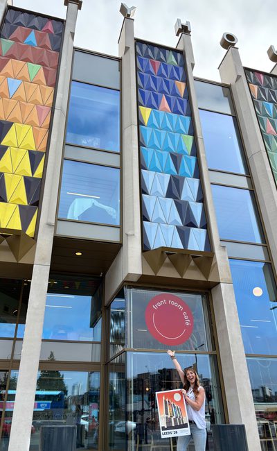 A woman with long dark wavy hair, wearing a white top and sunglasses on her head is standing in front of the iconic multicolored-triangle facade of Leeds Playhouse. She has one arm in the air, gesturing towards the building. In the other hand she’s holding an A2 poster, featuring a Bauhaus-style illustration of … Leeds Playhouse!