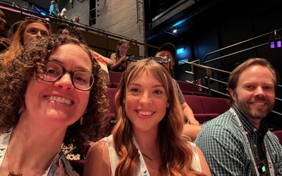 Three people sat in a theatre auditorium – a woman with a curly bob and glasses, another woman with long wavy dark hari, and a man with dark hair and a neat beard. All are smiling at the camera, as the first woman takes a selfie!