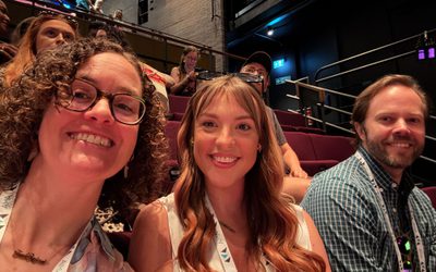 Three people sat in a theatre auditorium – a woman with a curly bob and glasses, another woman with long wavy dark hari, and a man with dark hair and a neat beard. All are smiling at the camera, as the first woman takes a selfie!