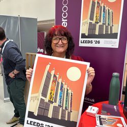 A smiling woman with a deep burgundy bob and fabulous large, dark-rimmed specs holds up an A2 poster depicting Leeds Playhouse, illustrated in a clay Bauhaus style.