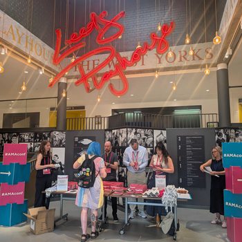 A large atrium, topped with a red neon sign reading 'Leeds Playhouse' in cursive lettering. Underneath, a conference registration desk with people getting their lanyards.