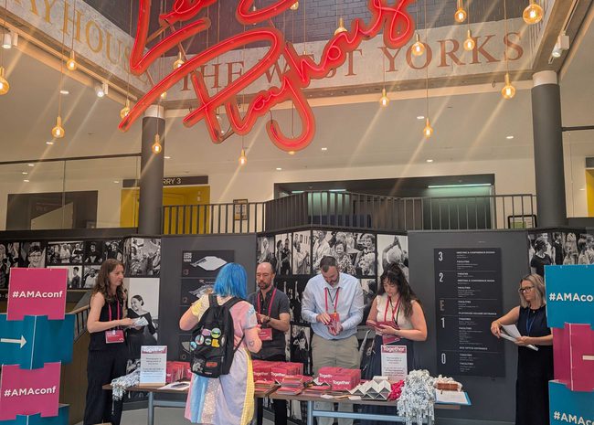 A large atrium, topped with a red neon sign reading 'Leeds Playhouse' in cursive lettering. Underneath, a conference registration desk with people getting their lanyards.