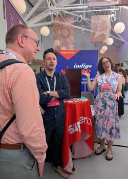 In a high-ceilinged room, several conference exhibition stands are set up. A smiling man stands close to the camera, talkign to another man next to him. A woman in the background smiles to camera and gestures towards the nearest conference stand.