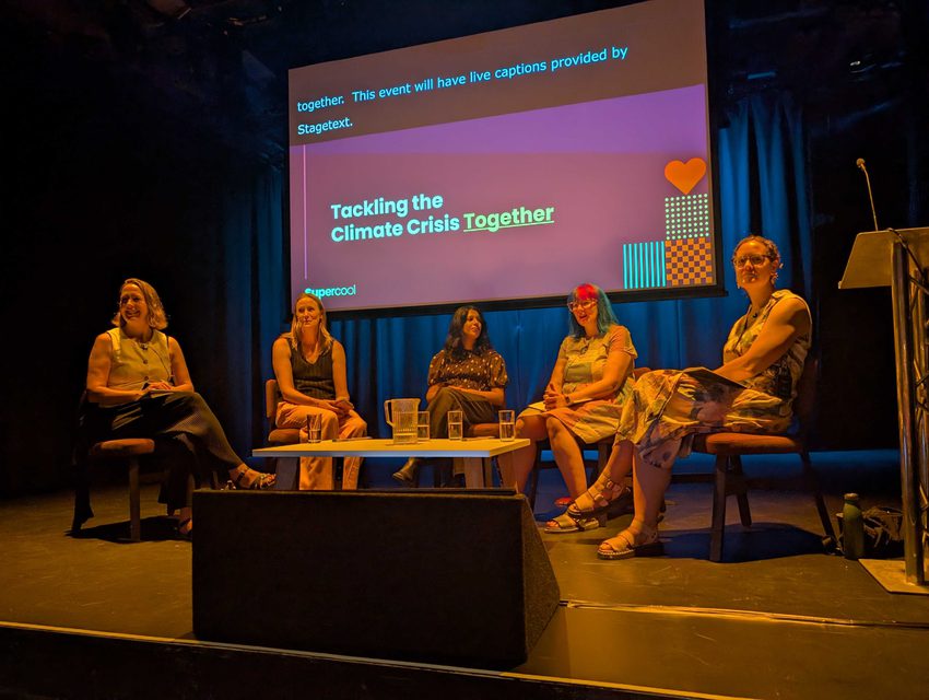 Five woman sat in a semi-cricle around a coffee table on stage at a conference, with a screen behind that reads "Tackling the climate crisis – together"