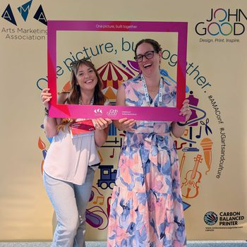 Two women laughing while holding a pink cardboard frame with the AMA logo on it