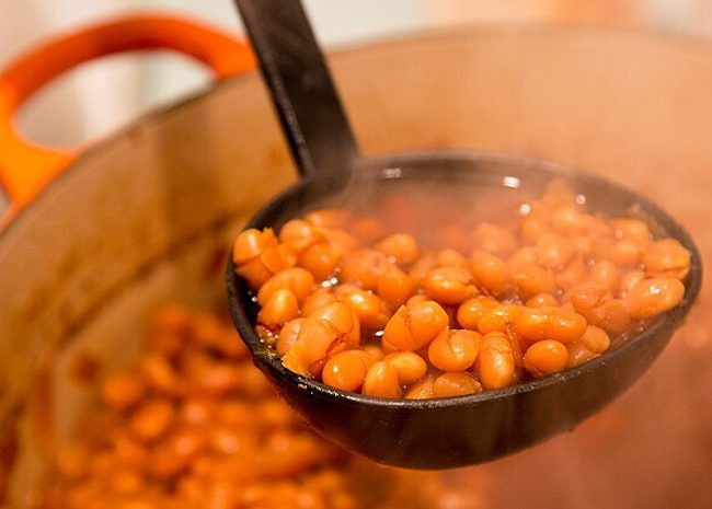 A black ladle holds a pile of steaming-hot baked beans. (Being lifted out of a huge orange pot full of baked beans)