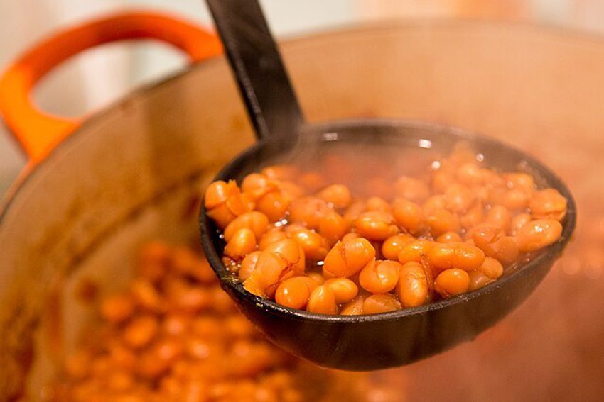 A black ladle holds a pile of steaming-hot baked beans. (Being lifted out of a huge orange pot full of baked beans)