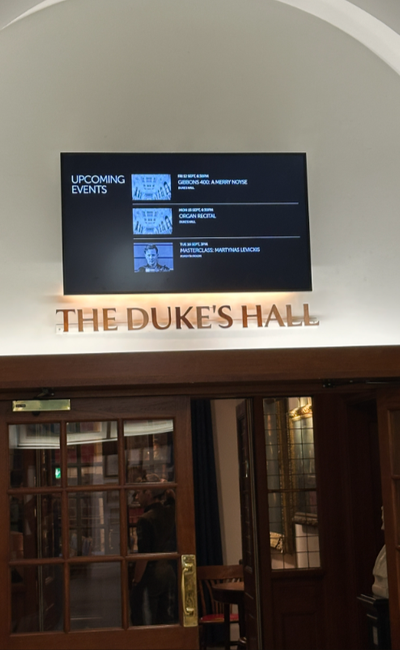 Above wooden-framed doorway, on a white wall with curved alcove above, and beneath it, signage reading "The Duke's Hall" is a venue screen listing upcoming events