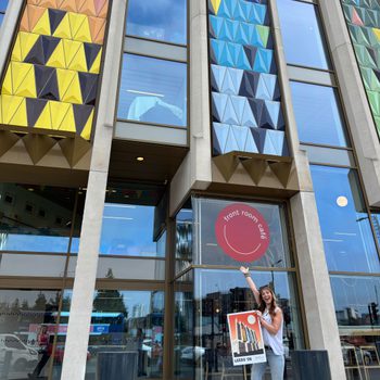 A woman with long dark wavy hair, wearing a white top and sunglasses on her head is standing in front of the iconic multicolored-triangle facade of Leeds Playhouse. She has one arm in the air, gesturing towards the building. In the other hand she’s holding an A2 poster, featuring a Bauhaus-style illustration of … Leeds Playhouse!