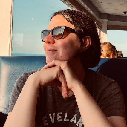 A light-skinned woman with a short burnette bob. She is wearing sunglasses, and has her elbows resting on a table in front of her as she looks to the left with the sun on her face. She's wearing a dark t-shirt with indecipherable letters on it.