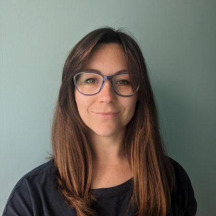 A light-skinned woman standing against a pale sage green wall. She has long brunette hair, and is wearing a dark collarless top, large, dark-rimmed glasses, and a gentle smile.