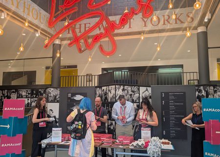 A large atrium, topped with a red neon sign reading 'Leeds Playhouse' in cursive lettering. Underneath, a conference registration desk with people getting their lanyards.