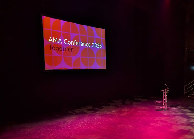 A dimly lit, red-tinged stage with an empty podium is backed-up by a red an purple presentation screen that reads "AMA Conference 2026 – Together".