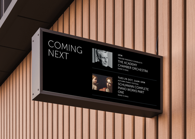 On an acousitc wooden-slat panelled wall, a black rectangular venue screen sticks out at 90 degrees. The screen reads "Coming next", and displays 2x event images, titles, dates and times